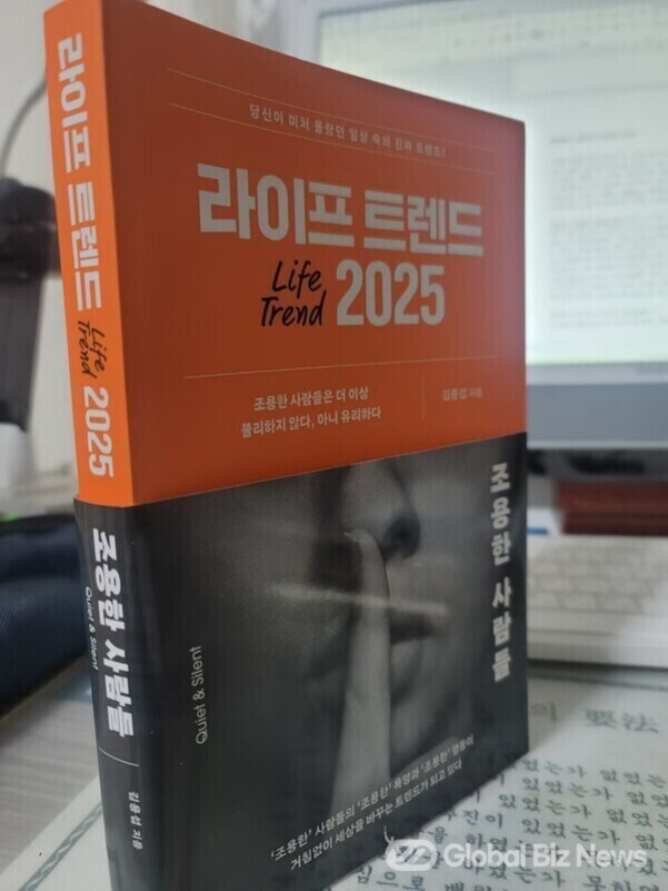'라이프 트렌드 2025'라는 제목의 책이 책상 위에 놓여 있고, 표지에는 조용히 하라는 제스처를 하는 사람의 흑백 사진이 있다. 배경에는 컴퓨터 모니터와 키보드가 보인다.