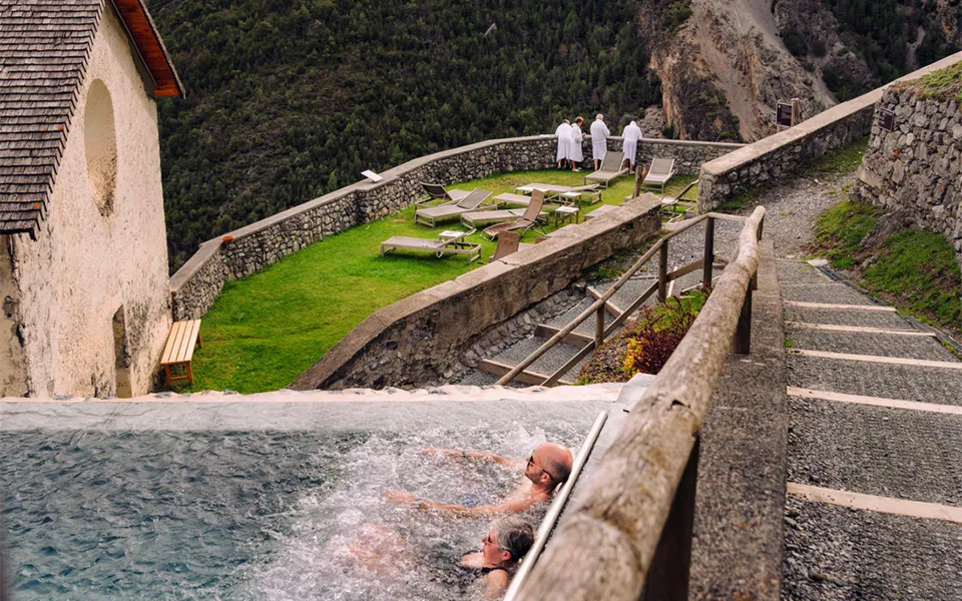 산속에 위치한 야외 온천탕에서 두 사람이 물에 들어가 있고, 위쪽 잔디밭에는 일광욕용 의자와 흰 가운을 입은 네 사람이 서 있다.