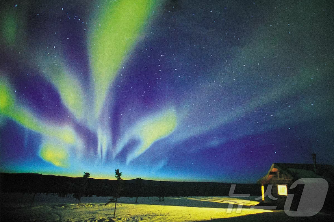 밤하늘에 펼쳐진 화려한 오로라와 별들, 눈 덮인 땅과 오른쪽에 작은 오두막이 보인다.